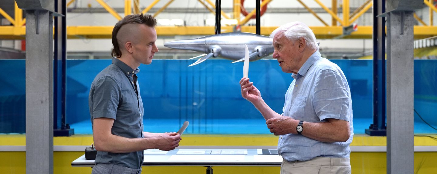 Luke Muscutt and Sir David standing facing each other in conversation. The robot hovers in the wave tank in the background.