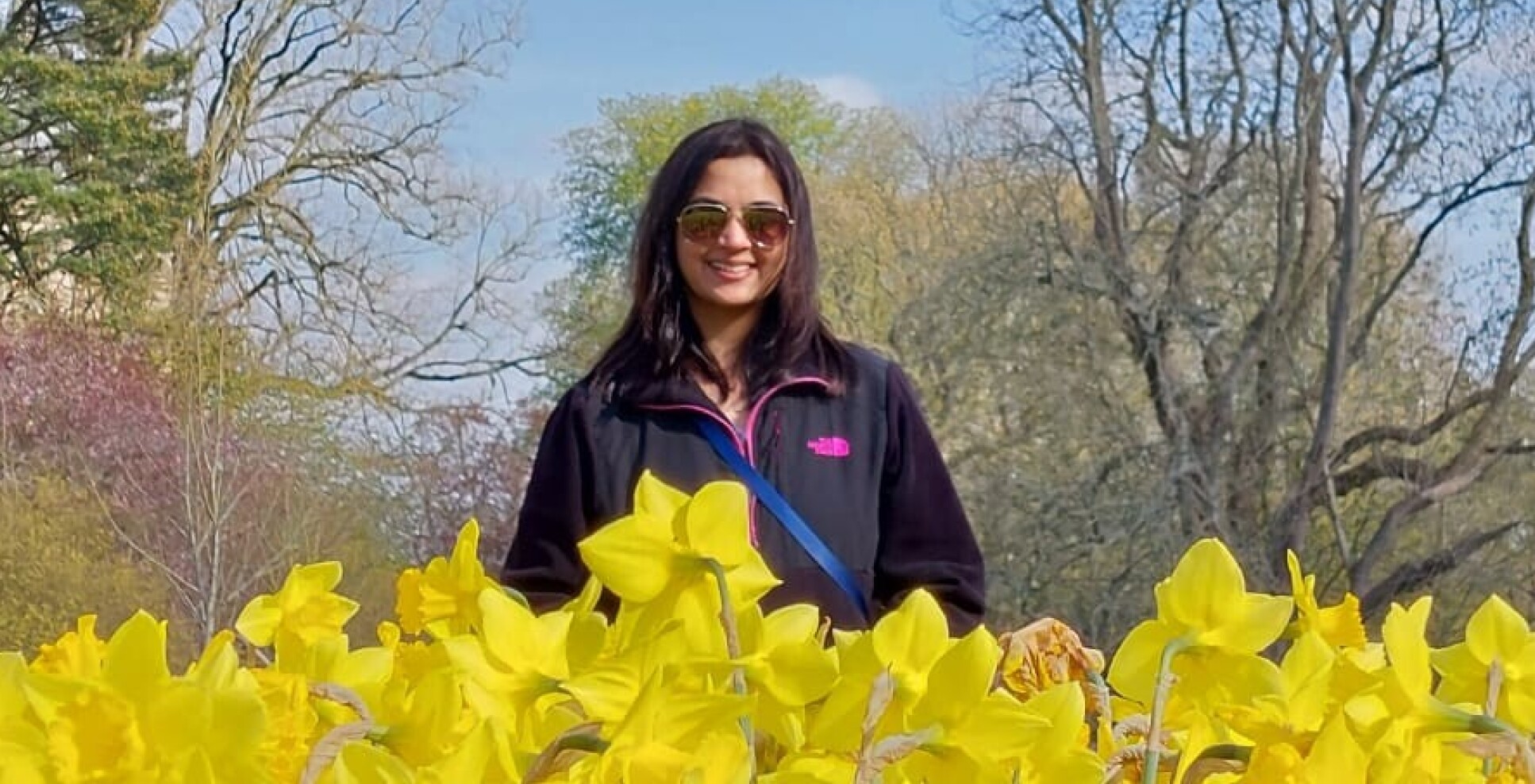 Dr Reshma Rao, a woman with brown hair, standing in a field of daffodils with trees and blue skies in the background i